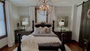 A bedroom with a large bed, a chandelier, a dresser, and a mirror. - Two Hearts Rambling Rose (Hermann)