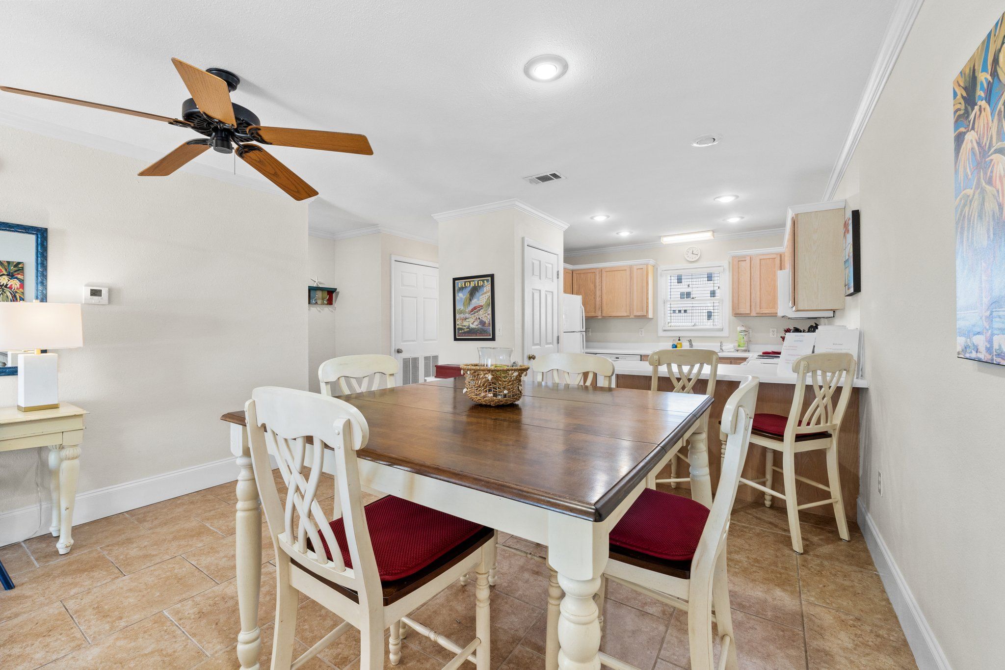 A dining area with a wooden table and white chairs, a ceiling fan, and a kitchen in the background.