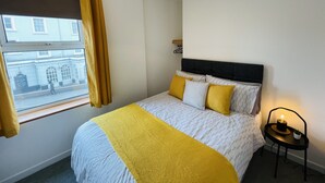 Une chambre à coucher avec un lit, une fenêtre avec des rideaux, une table de chevet avec une lampe, et une vue sur un bâtiment à l’extérieur.