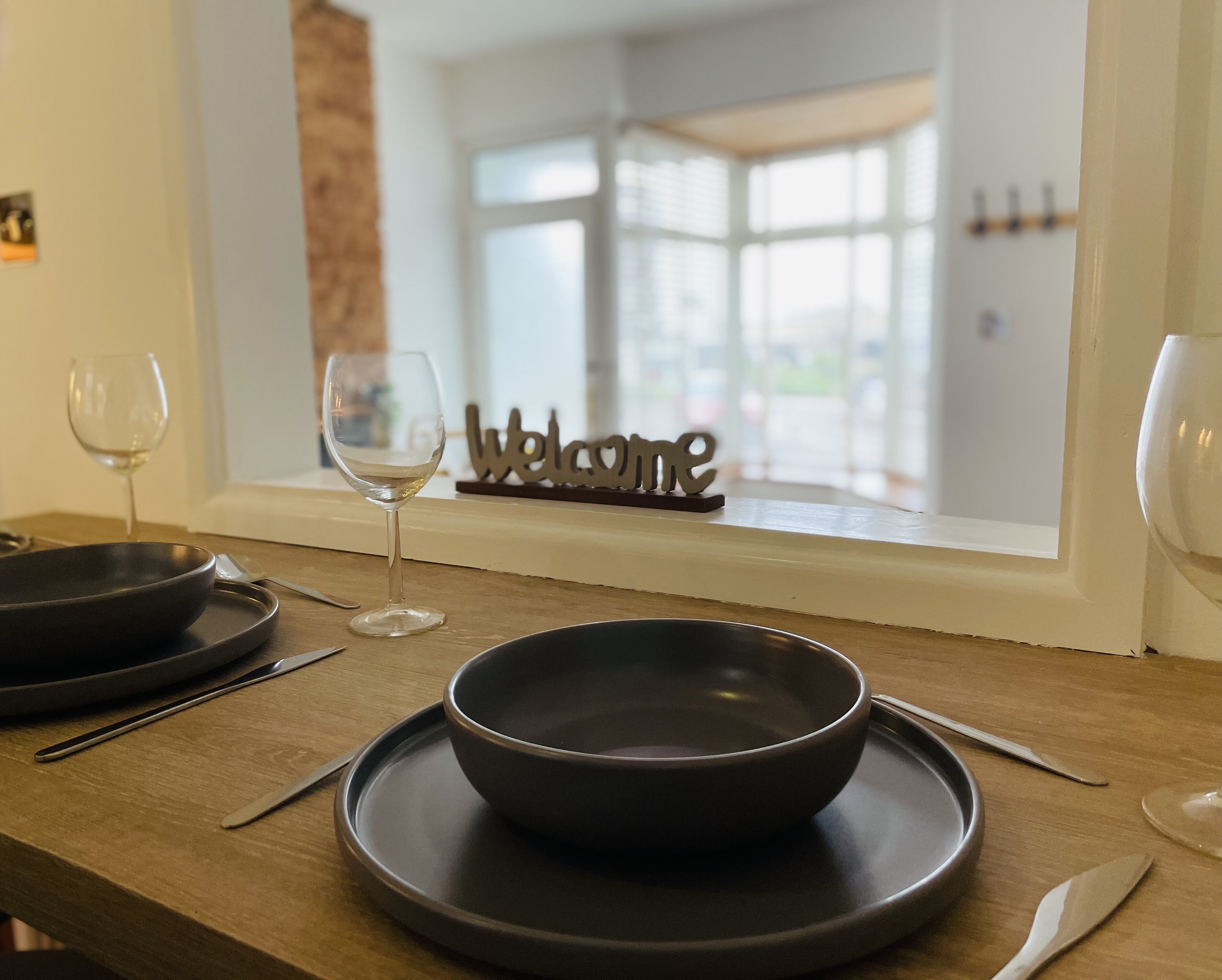 Une table à manger avec un saladier foncé, un verre à vin et des couverts. On peut voir une pancarte « Bienvenue » à travers une fenêtre.
