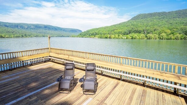 Eine Holzterrasse mit zwei Liegestühlen, die auf einen ruhigen See und bewaldete Hügel blicken.