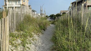 Un sendero arenoso bordeado por vallas de madera y hierbas altas, que conduce a propiedades frente a la playa con balcones.