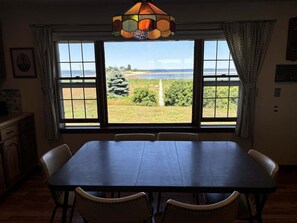 A dining room with a large table and chairs, a stained glass light fixture, and a view of a beach and greenery through large windows.