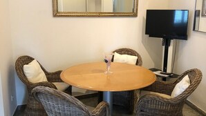 Ett rum med ett runt bord, fyra fåtöljer i rotting, en platt-TV och en stor spegel på väggen.