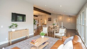 A spacious living area with a sofa, coffee table, and a television mounted on the wall. There is a staircase leading to an upper level, and a dining area with chairs and a table. - Lakefront Home! Game Tables and Boat Dock (Hendersonville)