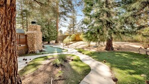Un patio trasero ajardinado con un muro de piedra, una terraza de madera, un camino de concreto y diversas especies de árboles.