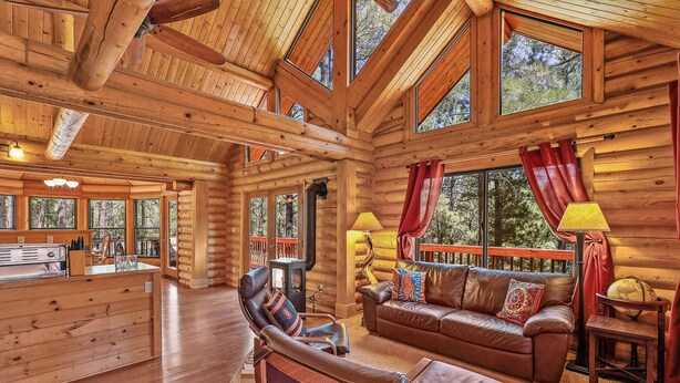 A log cabin interior with a wooden floor, a leather sofa, a dining area, and large windows offering views of the surrounding forest. - Kachina Tree House | 3BR | | Spend Spring in Flagstaff! (Flagstaff)