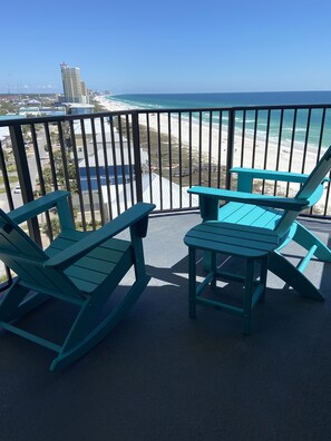 Ein Balkon mit zwei türkisblauen Adirondack-Stühlen und einem kleinen Tisch, der Blick auf einen Strand und den Ozean gewährt.