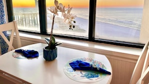 Ein Tisch mit einer Vase voller weißer Blumen, einer blauen Serviette und einem Teller mit Schildmotiven. Der Tisch steht in der Nähe eines Fensters mit Blick auf das Meer und einen Sonnenuntergang.