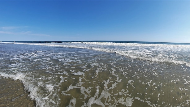 Ein Strand mit sanft anschwellenden Wellen, klarem Himmel und einem entfernten Steg.