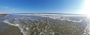 Una playa con olas suaves lamiendo la orilla, cielos despejados y un muelle a lo lejos.