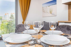 A dining table set with white plates and glasses, a view of buildings and greenery outside, and a painting on the wall.