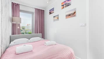 Un lit bien fait, recouvert d’une couette rose, avec des oreillers blancs et deux serviettes enroulées. Une fenêtre ornée de rideaux violets laisse entrer la lumière naturelle dans la pièce.