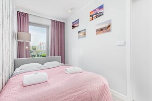 Ein ordentlich bezogenes Bett mit einer rosa Quilttdecke, weißen Kissen und zwei aufgerollten Handtüchern. Ein Fenster mit violetten Vorhängen lässt natürliches Licht in den Raum.