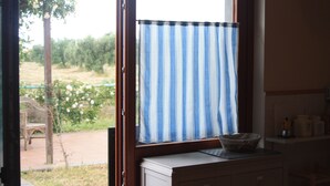 Een kamer met een raam, afgedekt met een blauw-wit gestreept gordijn, een houten bureau met een mand en een uitzicht op een tuin.