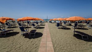 Een strand met rijen ligbedden en oranje parasols, een heldere lucht en een betegelde promenade.