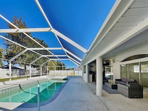 En pool utomhus med tak, vars takkonstruktion har ett geometriskt mönster, ett område med sittplatser bestående av en soffa och stolar, och en klarblå himmel.