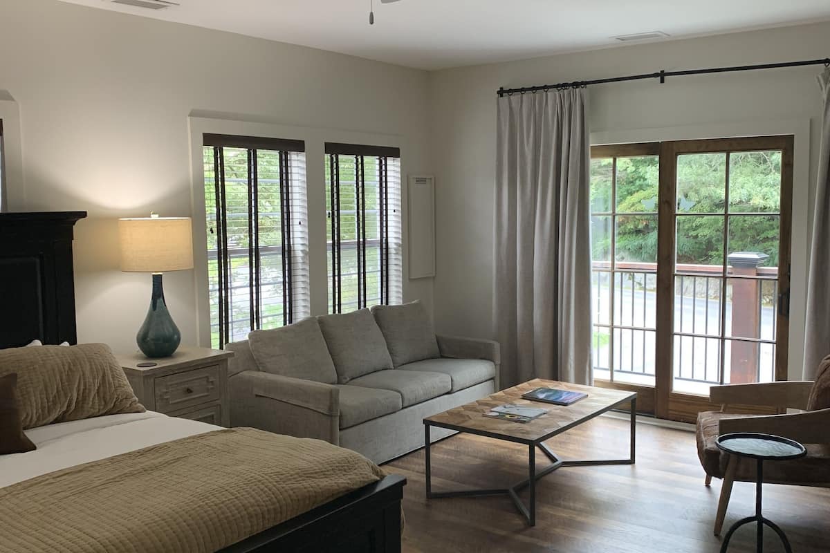 A spacious living room with a large bed, a sofa, a coffee table, and a ceiling fan. There are large windows with curtains and a view of greenery outside.