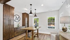 A kitchen with wooden cabinets, a dining table, pendant lights, and a view of greenery outside.