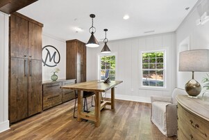 Una cocina con armarios de madera, una mesa de comedor, lámparas colgantes y vistas al exterior con vegetación.