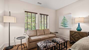 Uma sala de estar com um sofá, dois pufes, um abajur, uma mesinha lateral, um vaso decorativo e um quadro na parede.