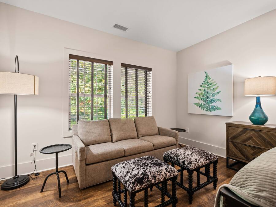 Uma sala de estar com um sofá, dois pufes, um abajur, uma mesinha lateral, um vaso decorativo e um quadro na parede.
