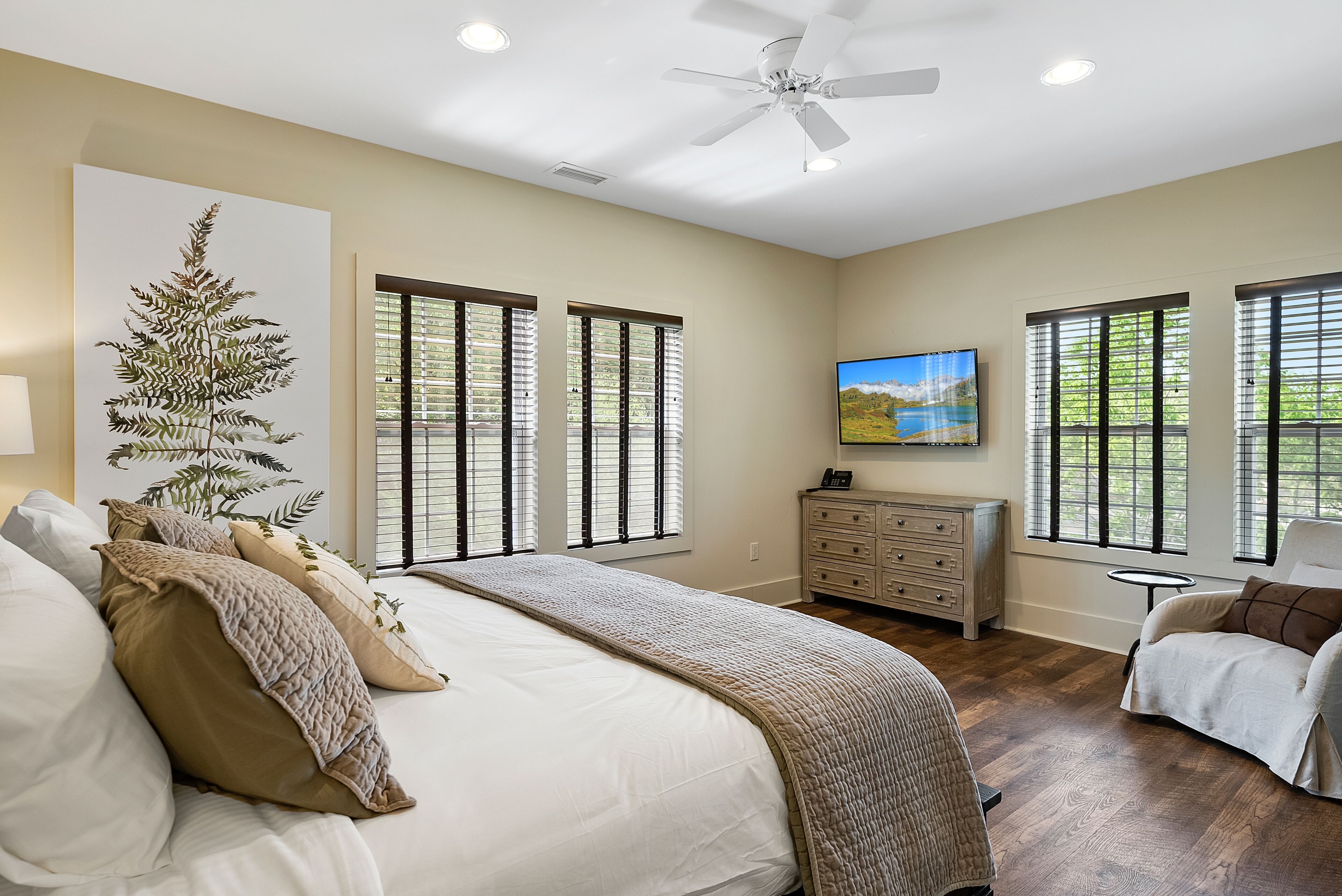 A bedroom with a large bed, a ceiling fan, a dresser, and a TV on the wall.