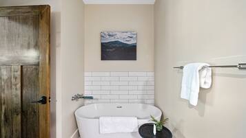 Un baño con una bañera, una puerta de madera, una toalla blanca, una mesita con una planta y un cuadro en la pared.