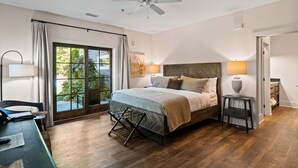 A bedroom with a bed, a desk, a chair, a ceiling fan, a window with curtains, and a view of greenery outside.