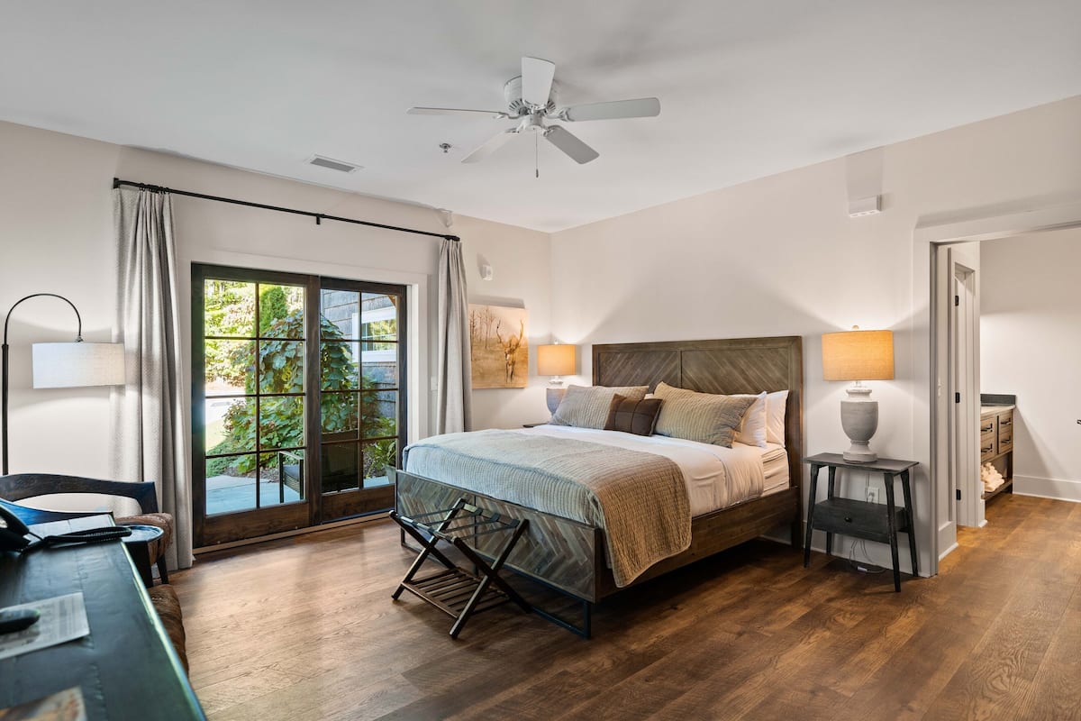 A bedroom with a bed, a desk, a chair, a ceiling fan, a window with curtains, and a view of greenery outside.