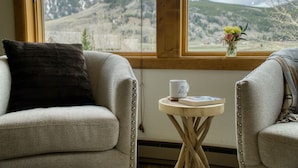A cozy room with two armchairs, a small wooden table with a book and a mug, and a window offering a mountain view.
