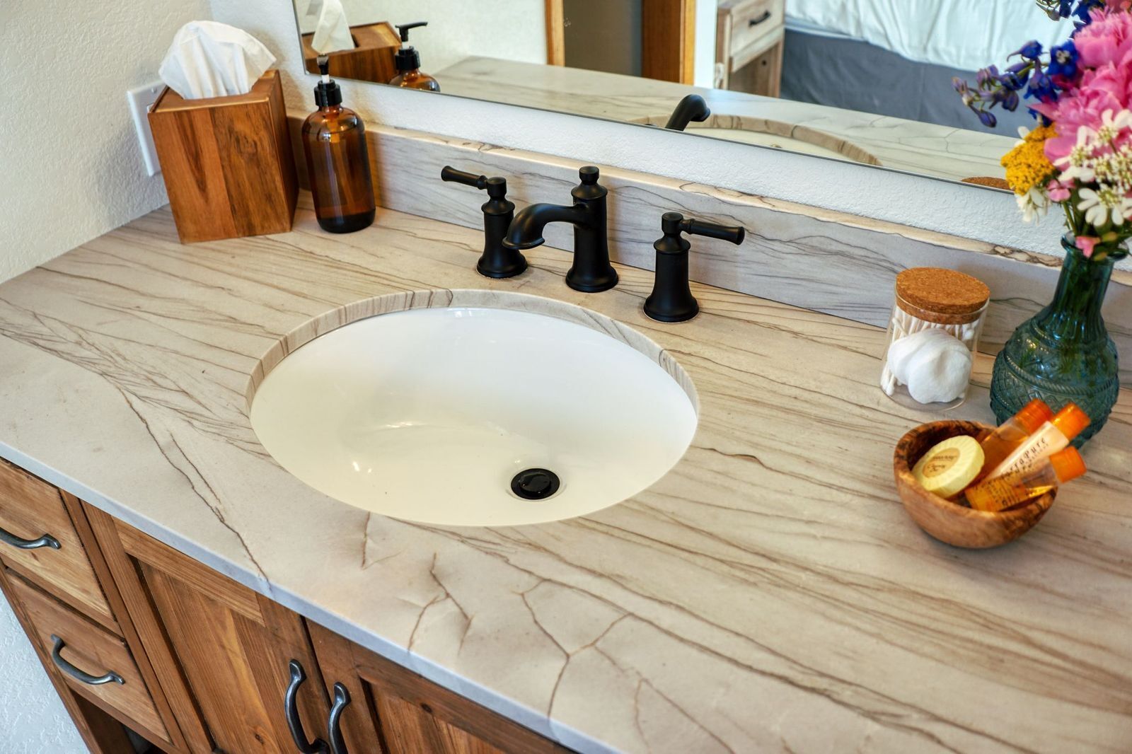 A bathroom with a marble sink, two black faucets, a wooden soap dish, a small wooden bowl with toiletries, a green glass bottle, and a jar with a cork lid.