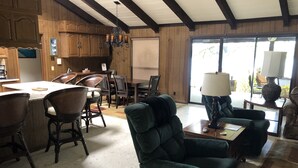 Uma sala ampla com uma poltrona verde grande, uma mesa de jantar com cadeiras, e uma cozinha com armários de madeira e um lustre.
