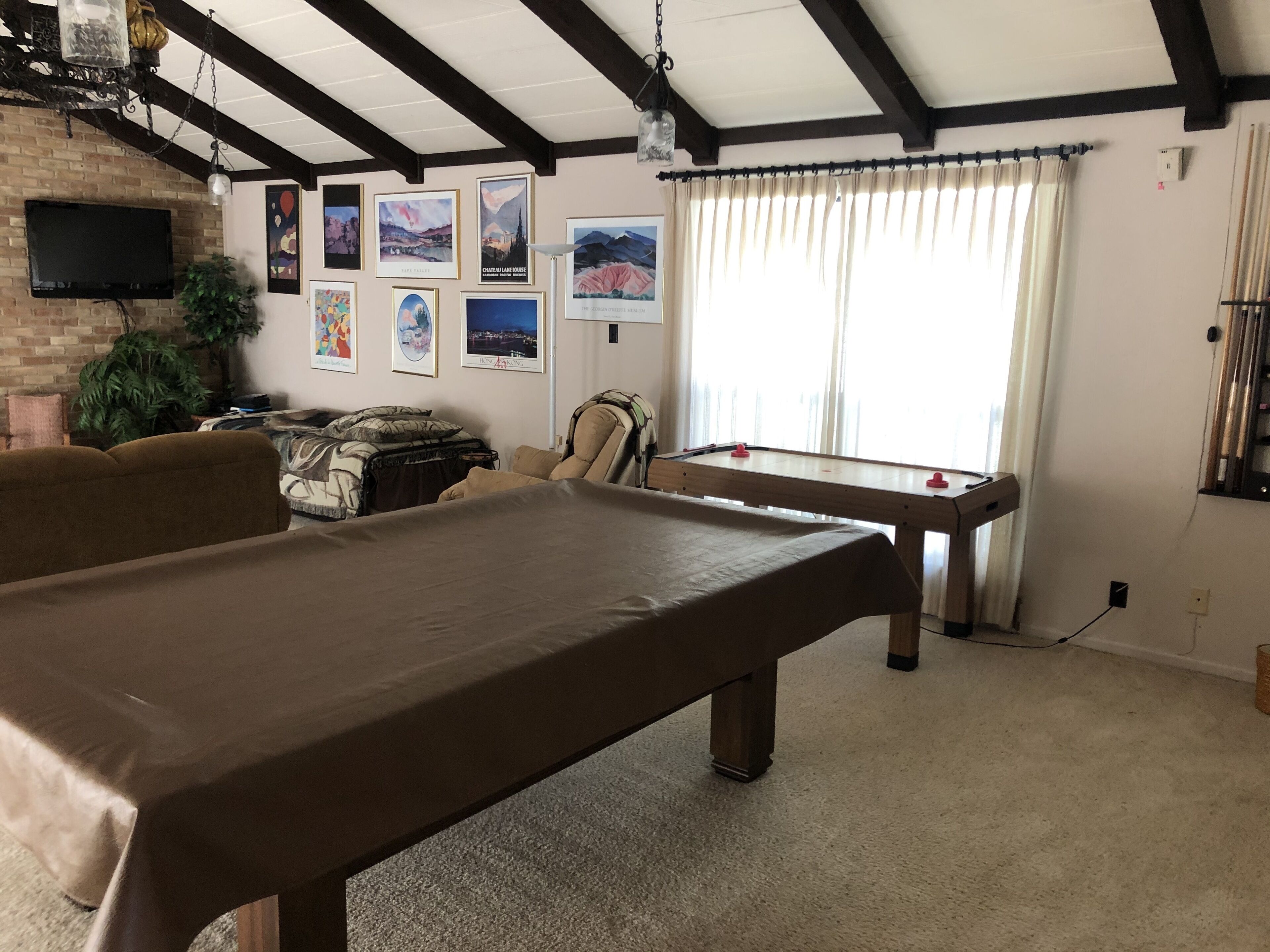 Una mesa de billar junto a una mesa de tenis, un sofá y un televisor instalado en una pared de ladrillo.
