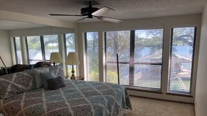 Quarto com cama grande, várias janelas, ventilador de teto e vista para um cais e para a água.
