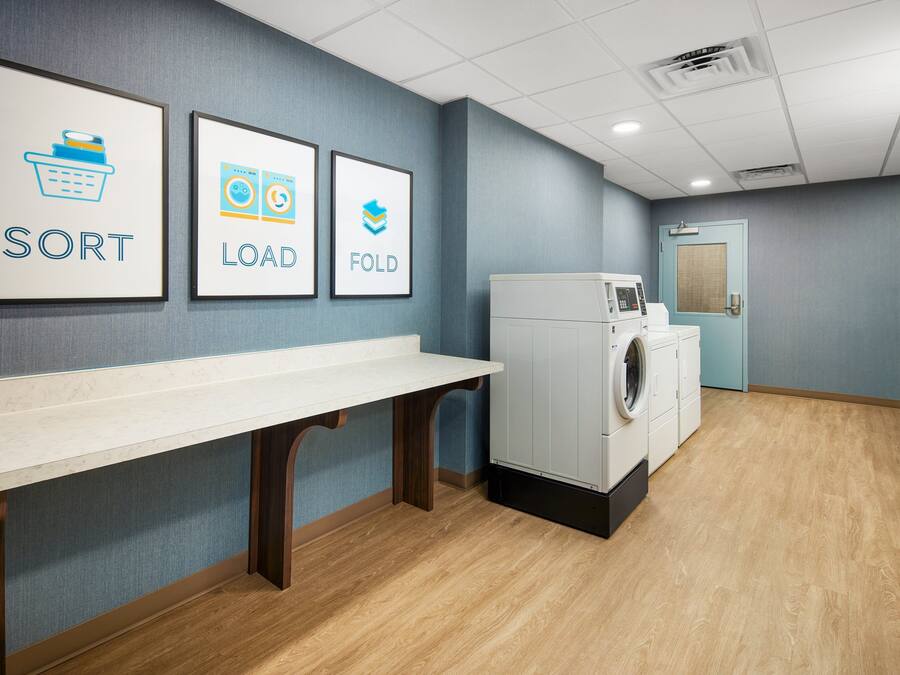 A laundry facility with a white counter, wall-mounted laundry machines, and framed instructions for sorting, loading, and folding.