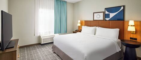A hotel room with a large bed, two bedside tables with lamps, a flat-screen TV on a wooden dresser, a window with curtains, and two framed pictures on the wall.