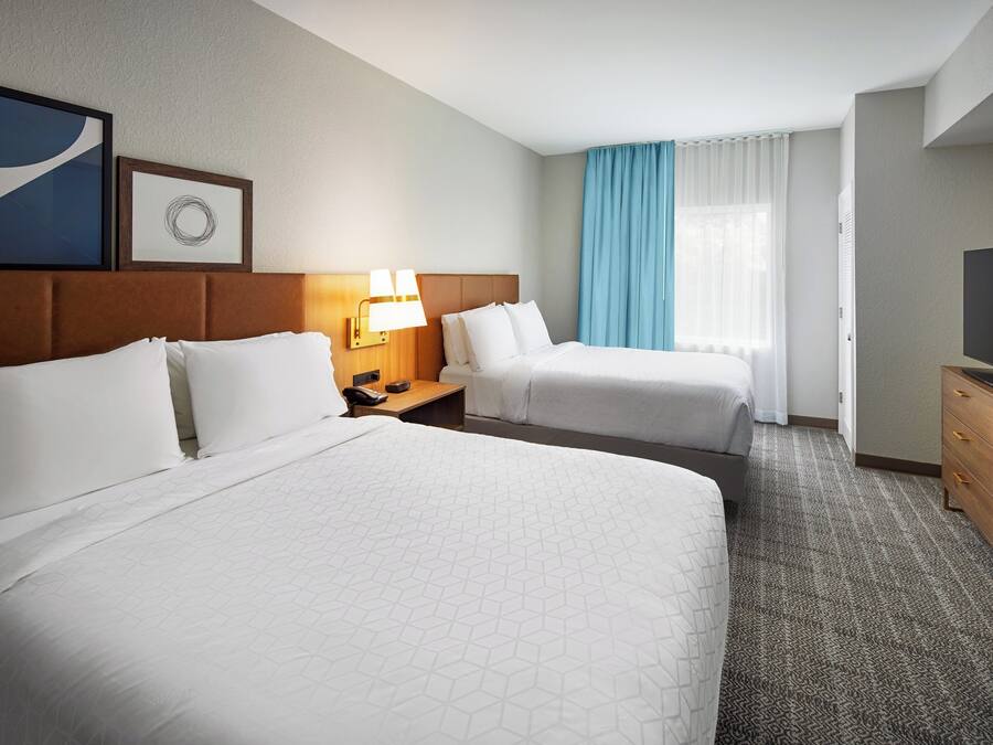 Ein Hotelzimmer mit zwei Betten, einem Schreibtisch, einem Fernseher und einem Fenster mit Vorhängen.