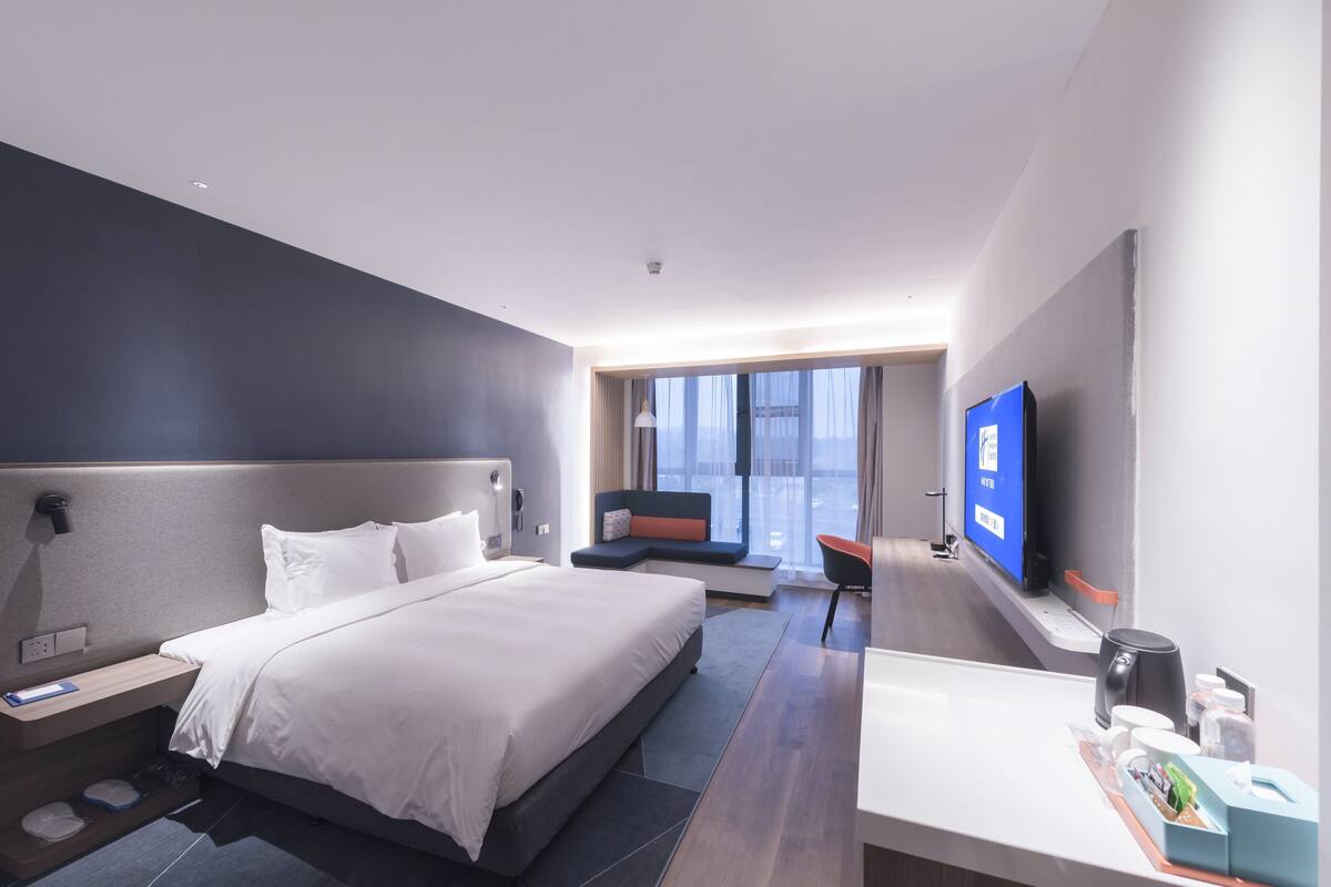 Habitación de hotel moderna con una cama grande, un escritorio, un televisor de pantalla plana y una zona de estar con vistas a la ciudad.
