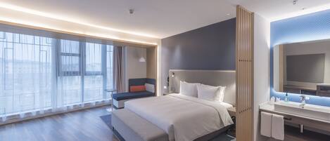 Een moderne hotelkamer met een groot bed, een zithoek, een badkamer met spiegel en grote ramen met uitzicht op de stad.