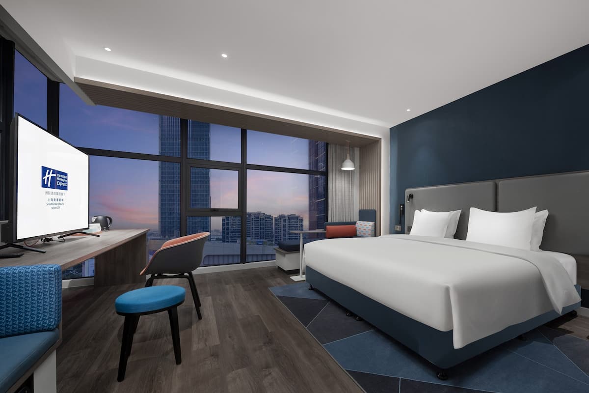 A modern hotel room with a large bed, a desk with a computer, and a view of the city skyline through large windows.