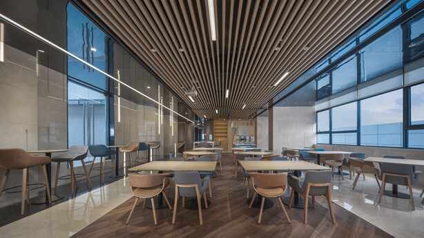 A modern dining area with wooden tables and chairs, large windows, and a slatted ceiling design.