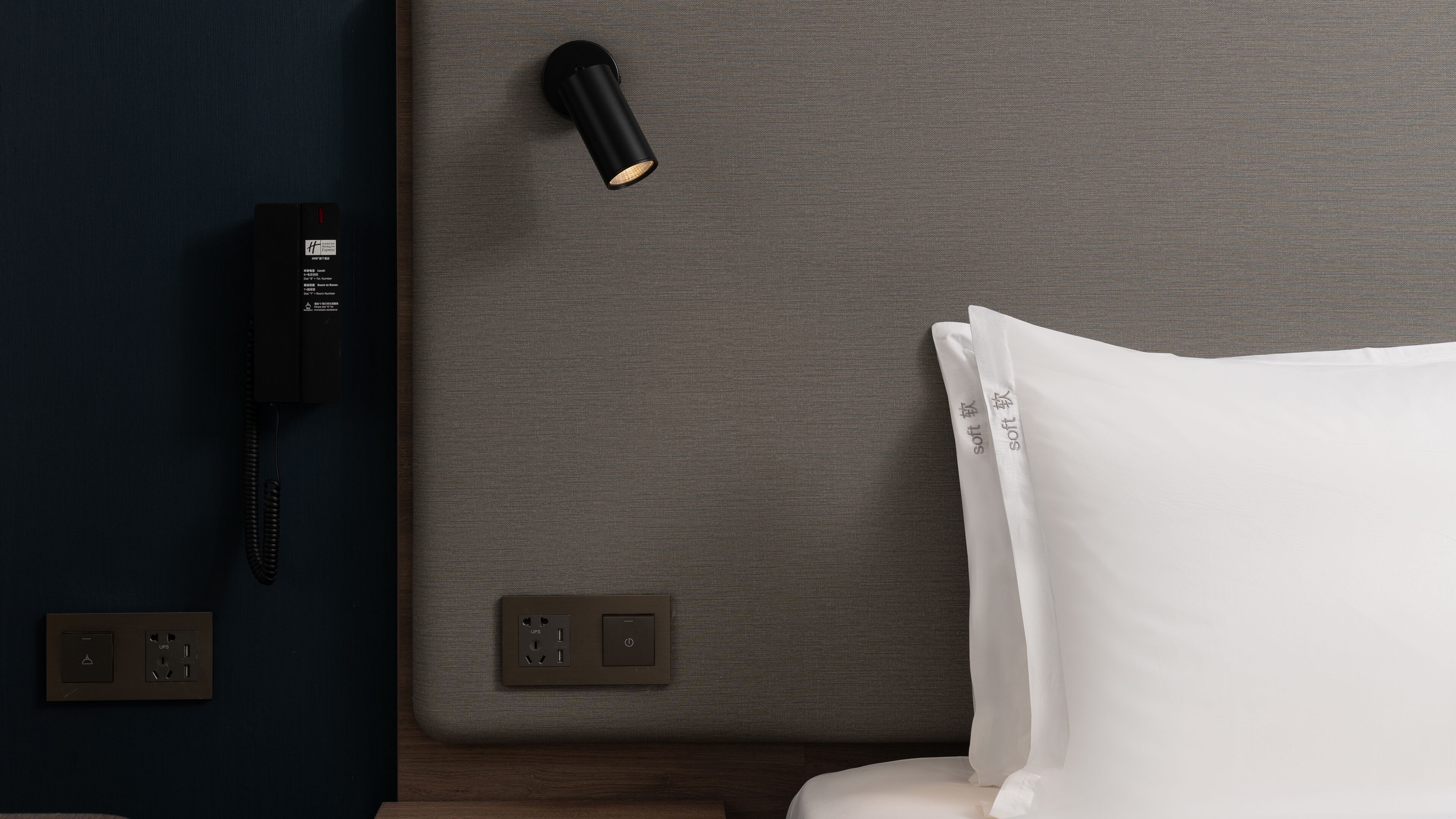 A hotel room with a bed, a bedside table, a lamp, a phone, and electrical outlets.
