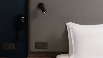 A hotel room with a bed, a bedside table, a lamp, a phone, and electrical outlets.