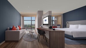 A modern hotel room with a large bed, a sofa, a desk with a TV, and a city view.