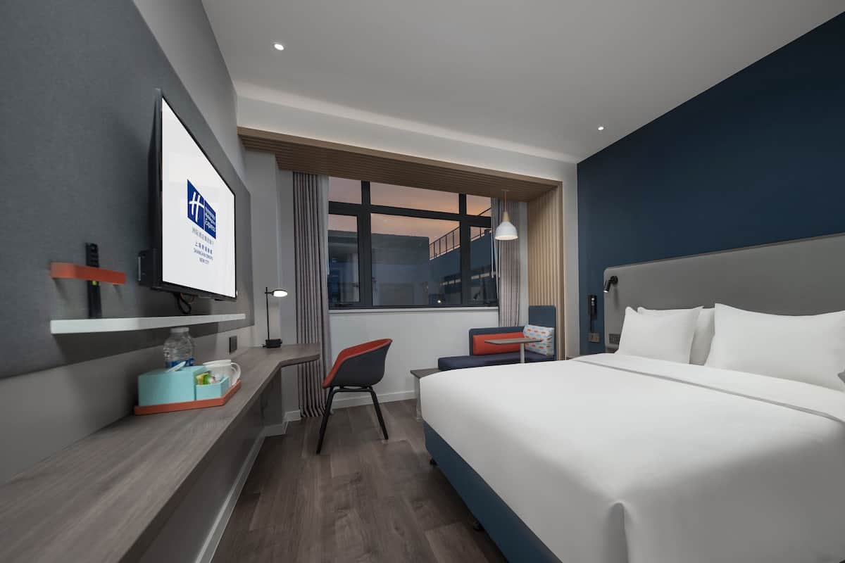 A modern hotel room with a large bed, a desk, a chair, and a TV on the wall.