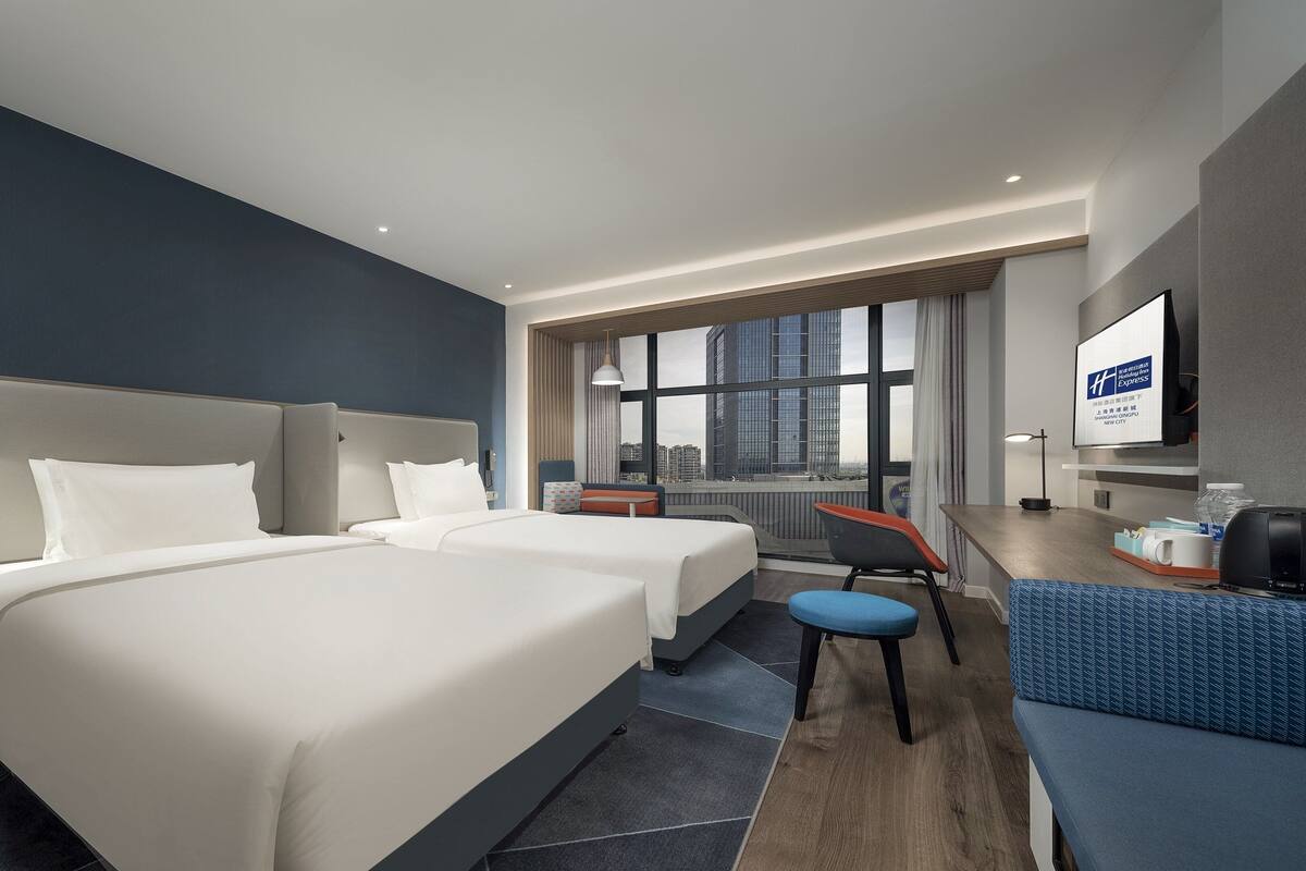 A hotel room with two beds, a desk, a chair, and a large window offering a city view.