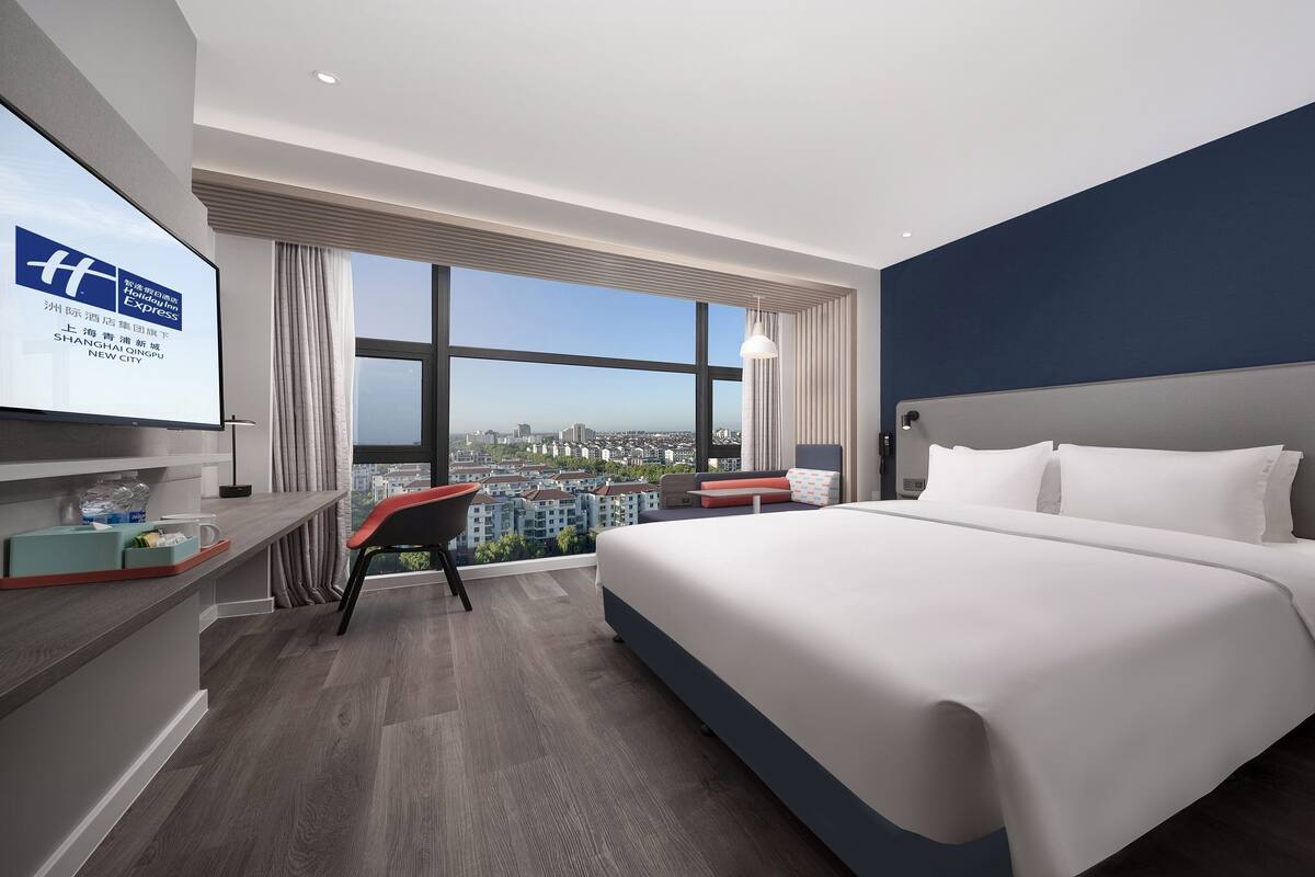 A modern hotel room with a large bed, a desk with a chair, and a large window offering a city view.