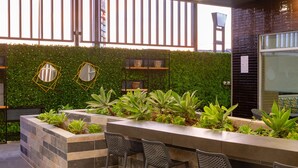 Un espace extérieur aménagé avec un banc et des chaises en pierre, entouré de plantes vertes et de miroirs décoratifs.