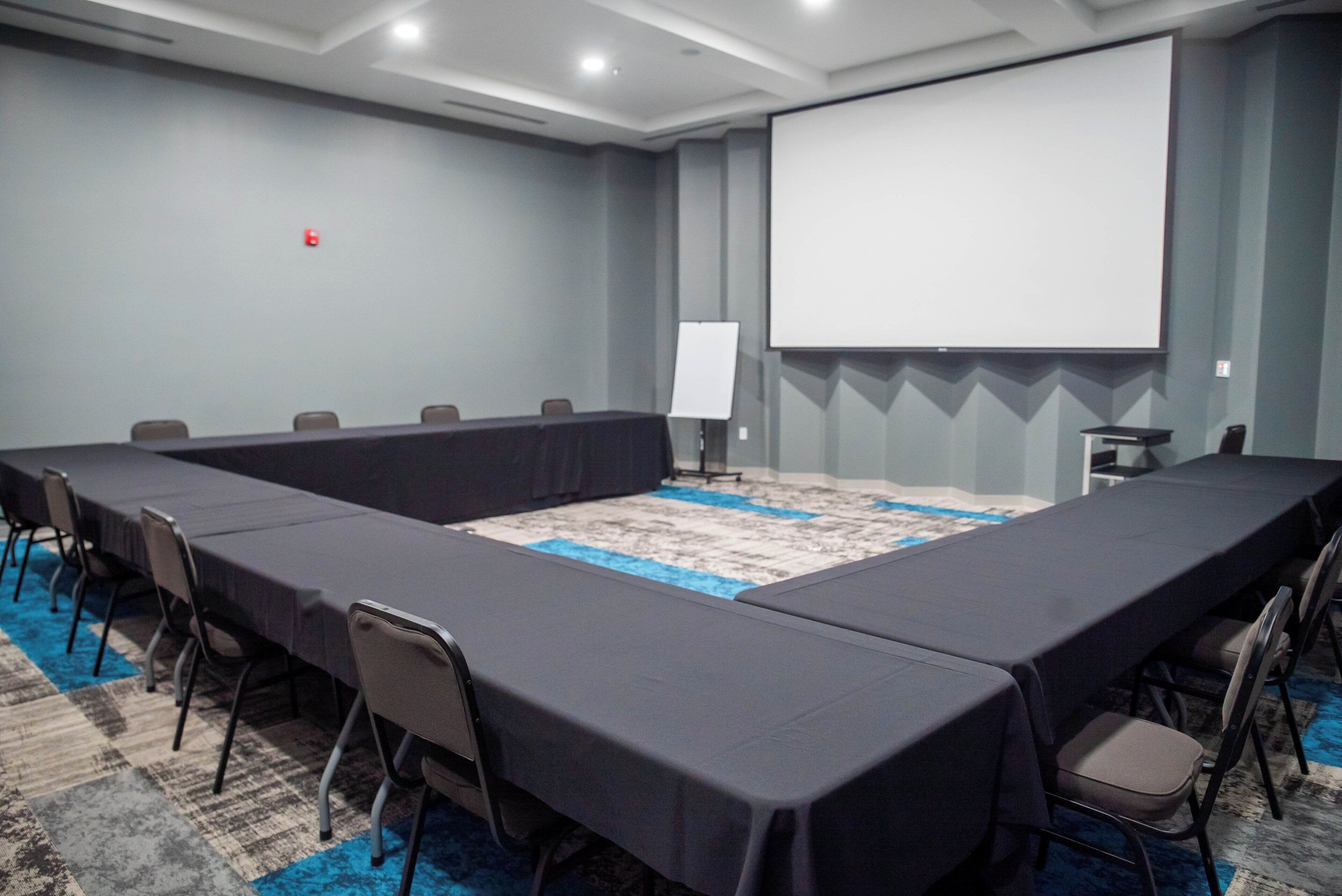 A conference room with a long table, chairs, a projector screen, and a carpeted floor.